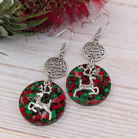 A pair of silver-toned dangle earrings with circular resin pendants featuring a red and green holiday pattern and a silver reindeer charm, displayed on a light wood surface with red flowers in the background.