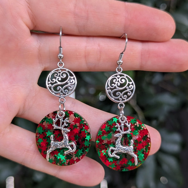 A pair of dangling earrings held in a hand. Each earring has a silver-tone filigree top and a round drop featuring a silver reindeer over a red and green glittery background.
