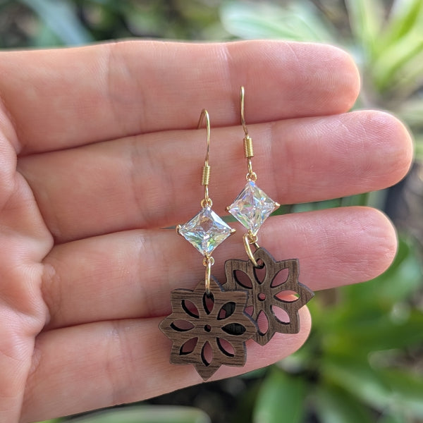 Plated gold earrings with crystals and wooden cut-out design held between fingers against a blurred green background.