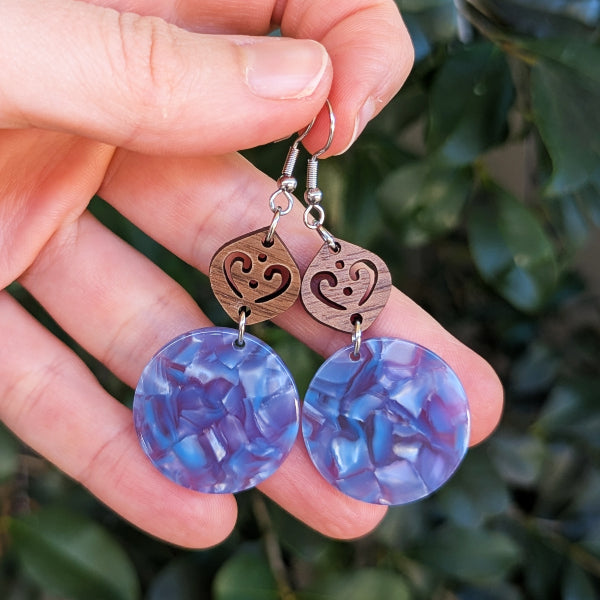 A pair of handmade dangle earrings held in a hand, featuring round marbled blue and purple resin pendants and wooden charms with hook wires, set against a blurred background of green leaves.