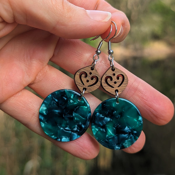 A close-up of a hand holding a pair of dangling earrings. Each earring has a silver fishhook ear wire, a small oval wooden charm with a heart-shaped cutout, and a large round green resin disc drop.