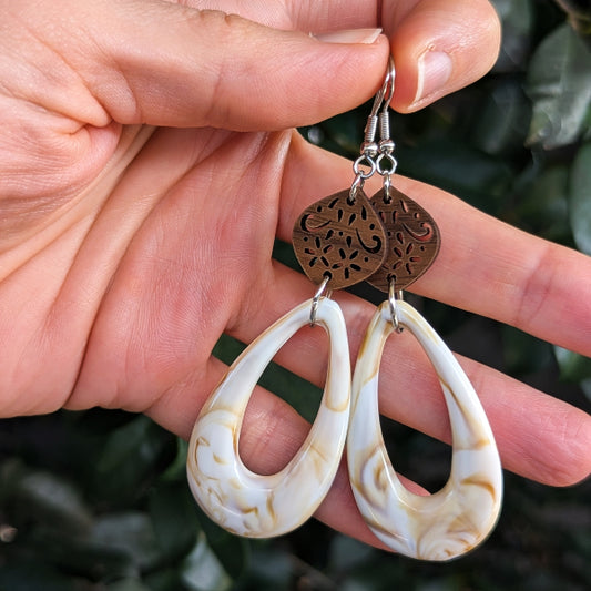A pair of handmade teardrop dangle earrings held in a hand. The earrings feature cream and brown marbled teardrop charms with cutout filigree wooden discs above them and silver hook ear wires.
