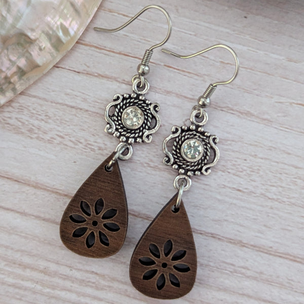Pair of earrings with wooden teardrop pendants and silver floral designs on a light wood surface.