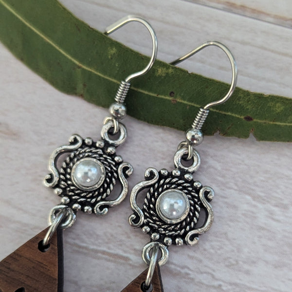 A pair of silver-toned bohemian dangle earrings with ornate filigree details and white faux pearl centers, resting on a green leaf over a light wood background.