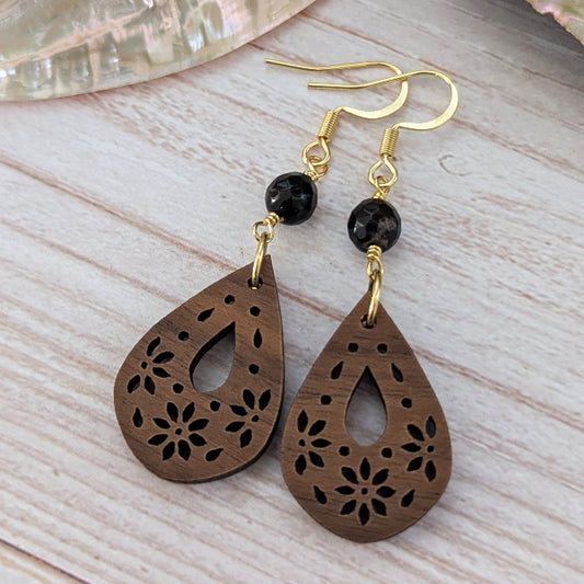 A pair of wooden teardrop dangle earrings with floral laser-cut patterns, gold-tone hooks, and black beads, displayed on a light wood background.