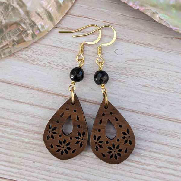 A pair of bohemian-style dangle earrings featuring laser-cut wooden teardrop pendants in a dark brown tone with floral cutouts, suspended from gold-tone hooks with a small black faceted bead, displayed on a light wood background.