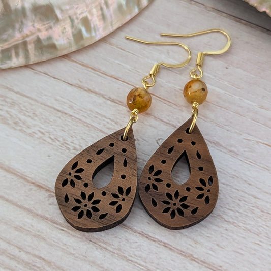 A pair of teardrop-shaped wooden dangle earrings with floral laser-cut patterns, gold-tone hooks, and a small amber bead, displayed on a light wood surface.