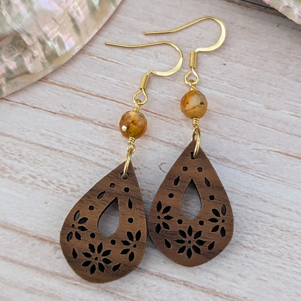 A pair of handmade dangle earrings featuring laser-cut wooden teardrop charms with floral patterns, a round amber bead, and gold-tone hook wires, displayed on a light wood background.