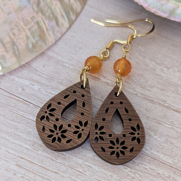 Wooden teardrop earrings with floral cutouts and orange beads on a wooden surface