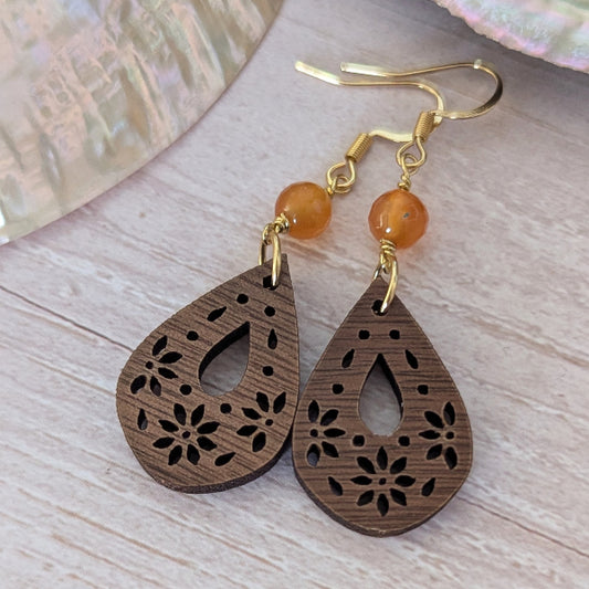 Wooden teardrop earrings with floral cutouts and orange beads on a wooden surface