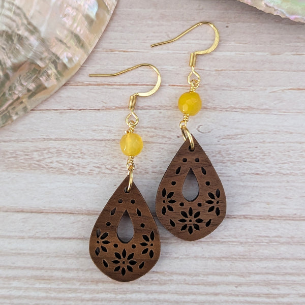 A pair of teardrop-shaped wooden dangle earrings with floral laser-cut patterns, gold hook wires, and a small round yellow bead, displayed on a light wood surface.