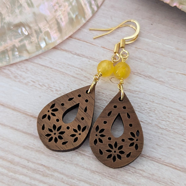 A pair of teardrop-shaped wooden dangle earrings with floral laser-cut patterns, yellow bead accents, and gold-tone hook ear wires, displayed on a light wood surface.