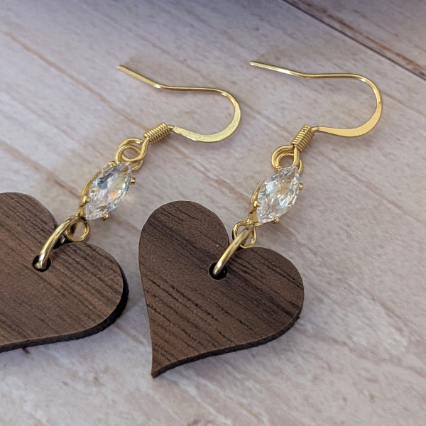 Heart-shaped wooden earrings with gold hooks and clear crystals on a light wooden surface.