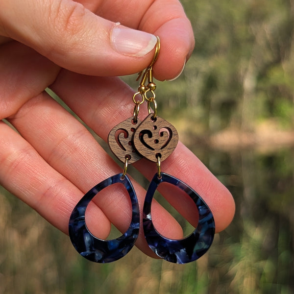A close-up of a hand holding a pair of dangling earrings against a blurred outdoor background. The earrings have gold hook wires, a small oval wooden charm with a cutout heart design, and large teardrop-shaped dark blue marbled resin drops.