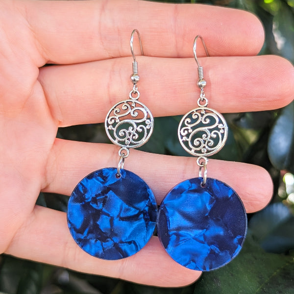 A pair of dangling earrings held in a hand, featuring silver-tone filigree circular tops and large marbled deep blue circular drop charms.
