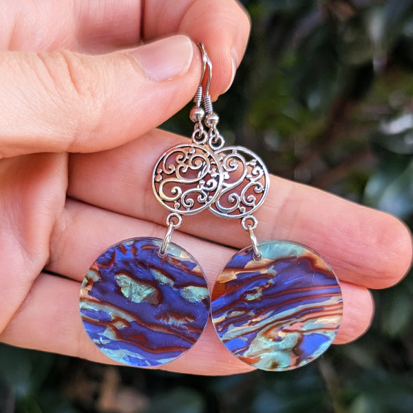 Silver earrings with colorful circular pendants held in a hand against a blurred green background