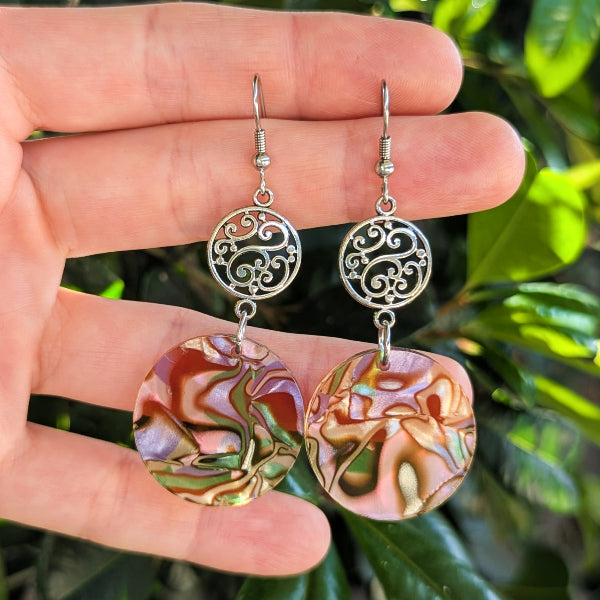 A pair of handmade dangle earrings held in a person's hand, featuring silver filigree circular links and round iridescent dangles with a marbled pink, green, and gold pattern, set against a background of green foliage.