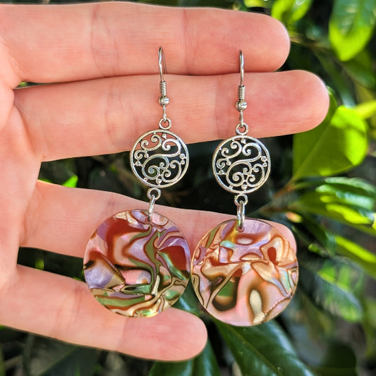 A pair of handmade dangle earrings held in a person's hand, featuring silver filigree circular links and round iridescent dangles with a marbled pink, green, and gold pattern, set against a background of green foliage.