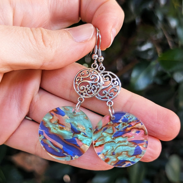 Hand holding a pair of earrings with intricate silver design and colorful circular drops.