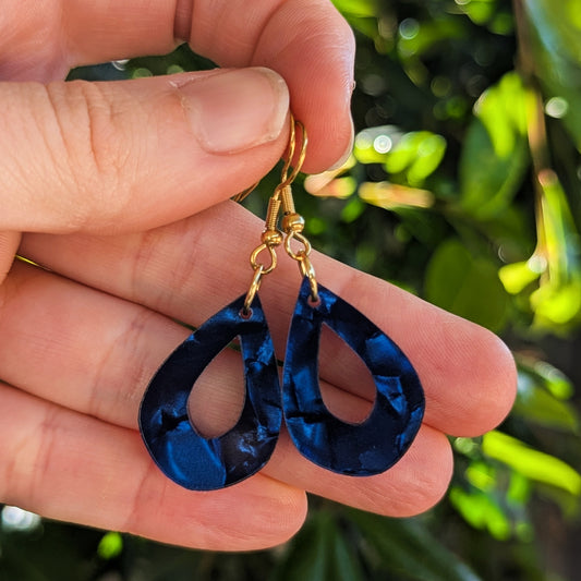 A close-up of a hand holding a pair of teardrop-shaped dangling earrings with gold hooks and deep blue marbled acrylic, set against a blurred green outdoor background.