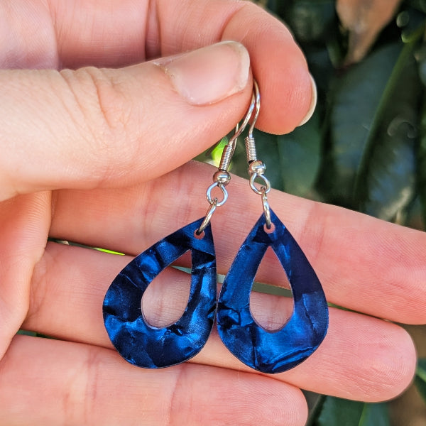 A pair of teardrop-shaped drop earrings with a marbled deep blue acrylic finish and silver hook ear wires, held in a person's hand against a blurred green background.