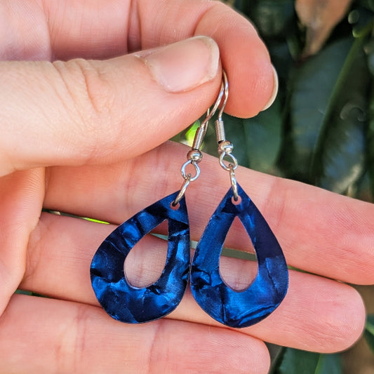 A pair of teardrop-shaped drop earrings with a marbled deep blue acrylic finish and silver hook ear wires, held in a person's hand against a blurred green background.