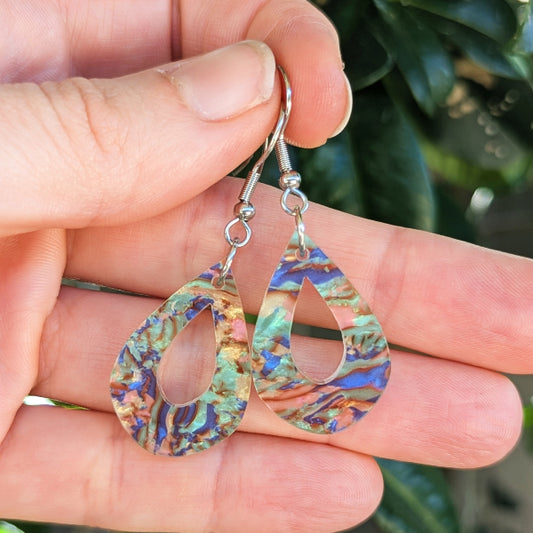 A pair of teardrop-shaped dangle earrings with iridescent acrylic and silver hook wires, held in a person's hand with a blurred green outdoor background.