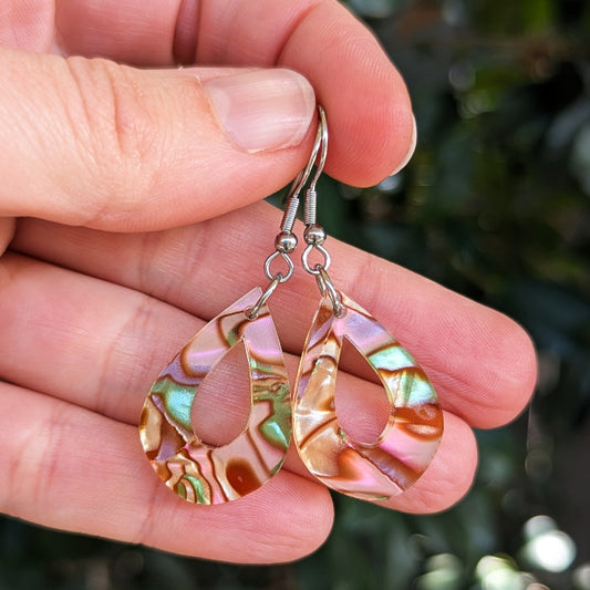 A close-up of a hand holding a pair of teardrop-shaped earrings made from iridescent acrylic with silver fishhook wires, set against a blurred green outdoor background.
