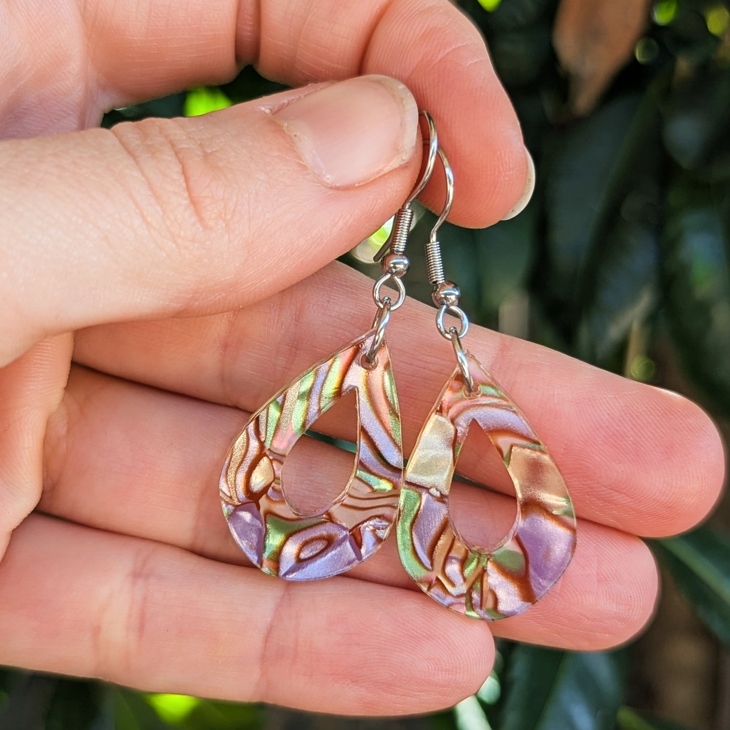 A pair of teardrop-shaped dangle earrings with iridescent acrylic and silver hook ear wires, held in a person's hand against a blurred green outdoor background.