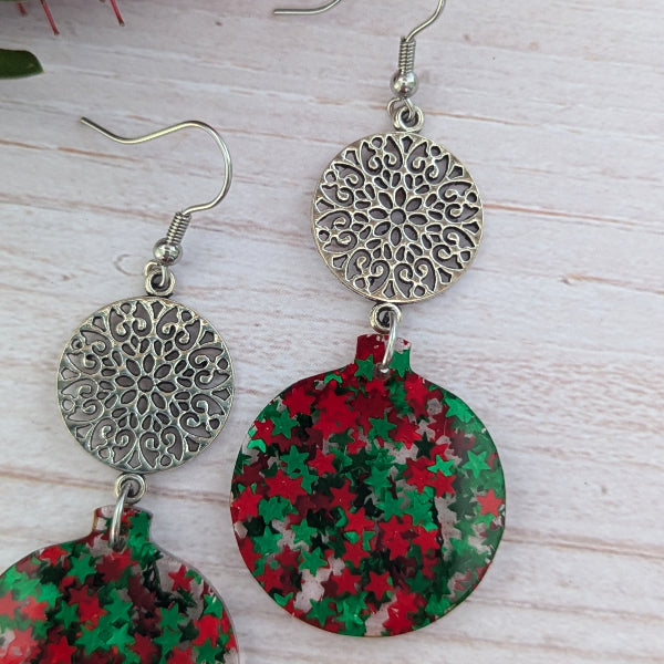 A pair of dangling earrings with ornate silver filigree circular tops and round lower charms covered in red and green star confetti, displayed on a light wood background.