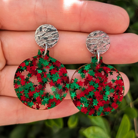A pair of round drop earrings with textured silver studs and circular dangles covered in red and green star-shaped glitter, shown resting on a person's hand against a blurred green background.