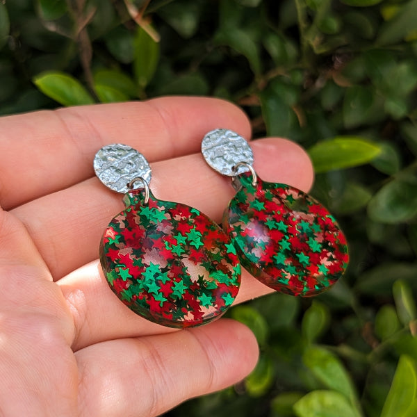 A pair of dangling earrings with textured silver studs and oval drops filled with red and green star-shaped glitter, held in a person's hand against a blurred green leaf background.