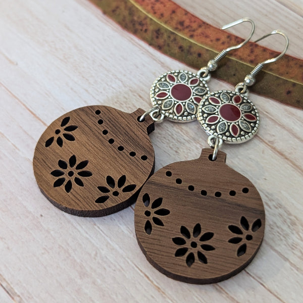 A pair of handmade wooden ornament dangle earrings with laser-cut floral patterns, featuring silver-toned filigree circular charms with red inlay, displayed on a light wood surface.