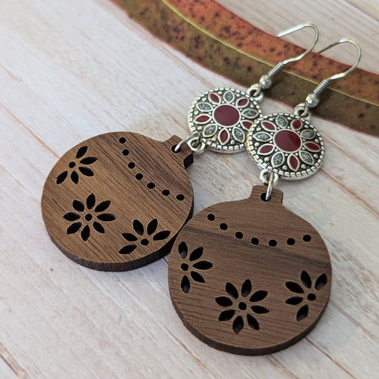 A pair of handmade wooden ornament dangle earrings with laser-cut floral patterns, featuring silver-toned filigree circular charms with red inlay, displayed on a light wood surface.