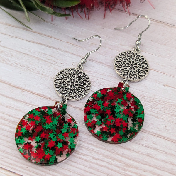 A pair of dangling earrings with silver-tone filigree round hooks and circular dangles covered in red and green star-shaped glitter, displayed on a light wood surface.