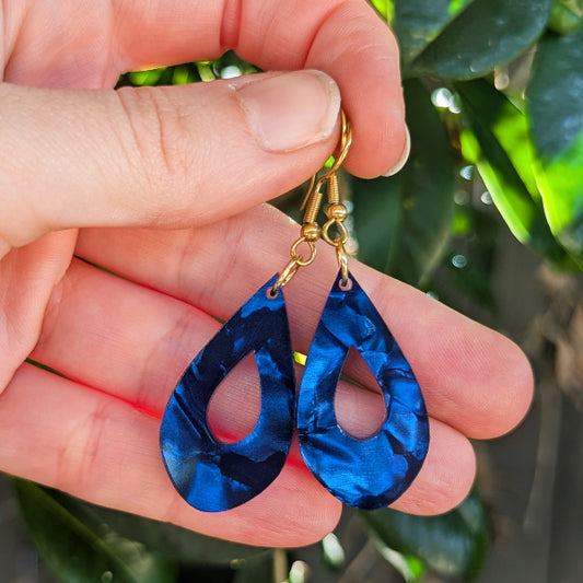 A pair of teardrop-shaped blue marbled earrings with gold hook ear wires, held in a person's hand against a background of green leaves.