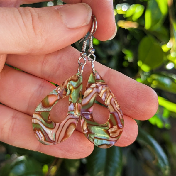 A pair of teardrop-shaped dangle earrings with iridescent acrylic inlays showing green, gold, and pink patterns, held in a person's hand against a blurred background of green foliage.