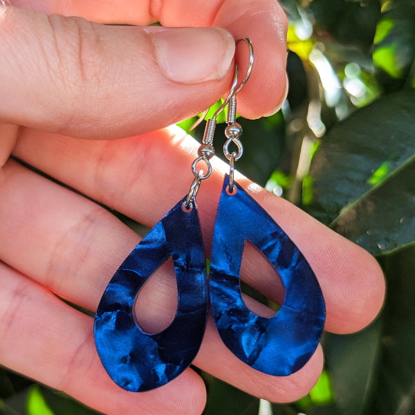 A pair of teardrop-shaped dangling earrings with a glossy blue marbled pattern, held by a hand against a background of green leaves.