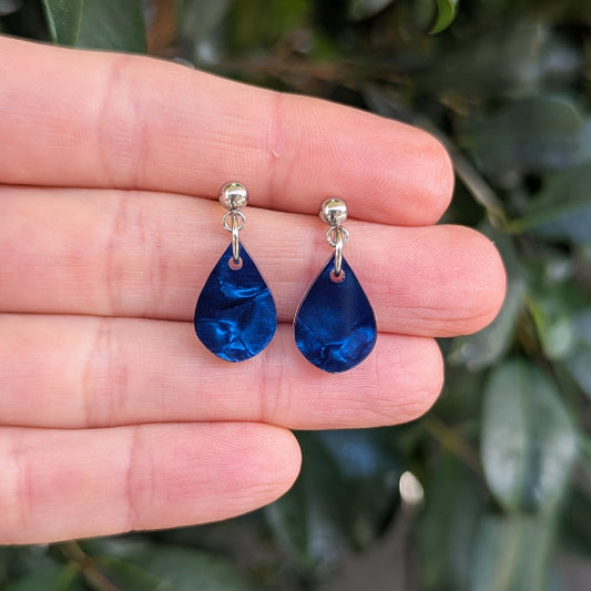 A pair of deep blue teardrop dangle earrings with silver posts, held on a person's fingers with a blurred background of green leaves.