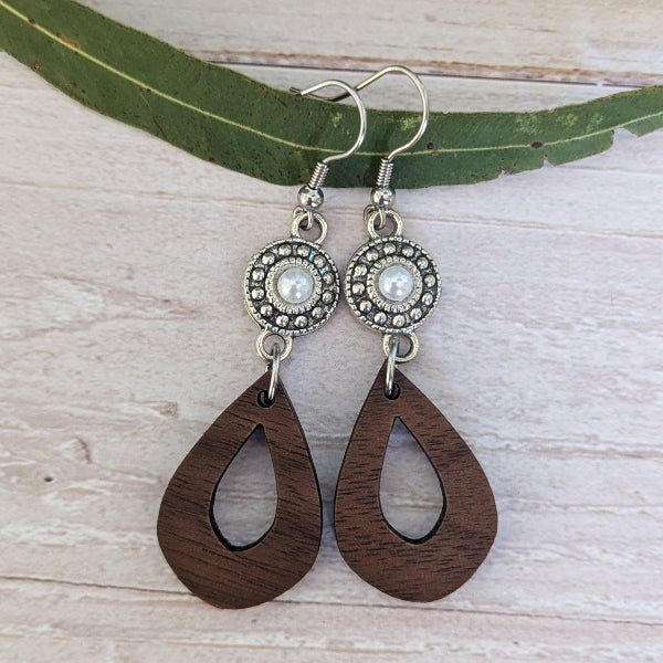A pair of handmade teardrop dangle earrings with dark brown wooden pendants and silver-toned metal hoops, displayed on a green leaf over a light wood background.