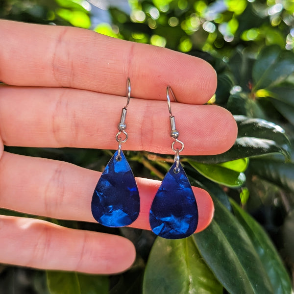 A pair of teardrop-shaped blue acrylic earrings with silver hook wires, held between a person's fingers against a background of green leaves.