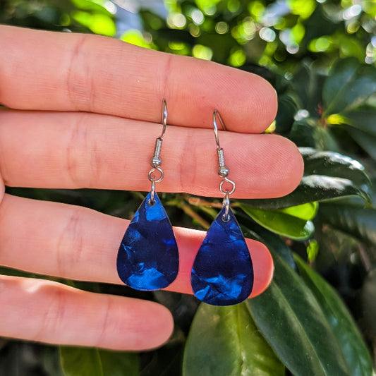 A pair of teardrop-shaped blue acrylic earrings with silver hook wires, held between a person's fingers against a background of green leaves.