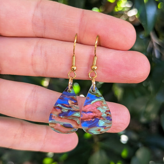A pair of teardrop-shaped dangle earrings with gold hook wires and marbled iridescent acrylic drops, held between a person's fingers against a blurred green outdoor background.