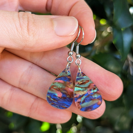 A close-up of a hand holding a pair of teardrop-shaped dangling earrings with iridescent blue, green, and pink marbled acrylic pendants and silver hook wires, against a blurred green outdoor background.