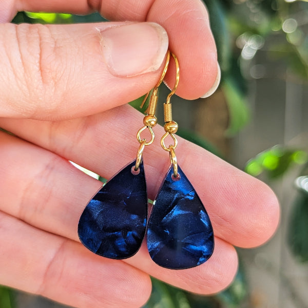 A pair of teardrop-shaped dark blue marbled earrings with gold hook wires, held in a person's hand against a blurred green outdoor background.