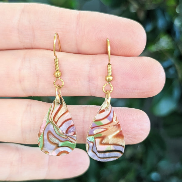 A pair of gold-tone teardrop dangle earrings with marbled iridescent acrylic drops, held between fingers against a blurred green outdoor background.