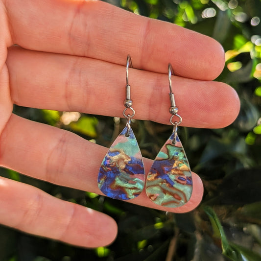 A pair of teardrop-shaped dangle earrings with iridescent blue, green, and gold marbled acrylic pendants on silver hook wires, held in a person's hand against a blurred green outdoor background.