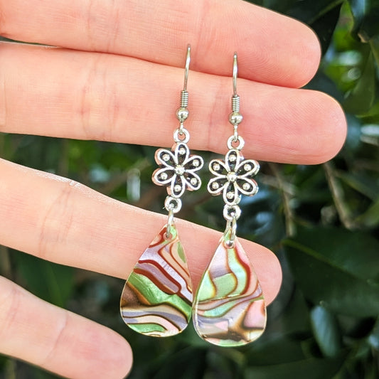 A pair of silver-tone dangle earrings with floral cutout charms and teardrop-shaped iridescent drops, held between fingers against a blurred green outdoor background.