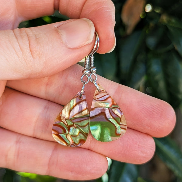 A pair of teardrop-shaped dangle earrings with iridescent gold, green, and copper marbled acrylic pendants on silver hook wires, held in a person's hand against a blurred green background.