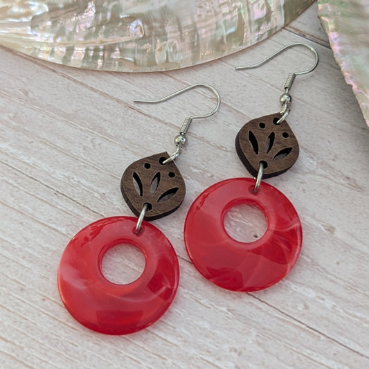 Pair of earrings with red circular pendants and brown leaf-like connectors on a light wooden surface.